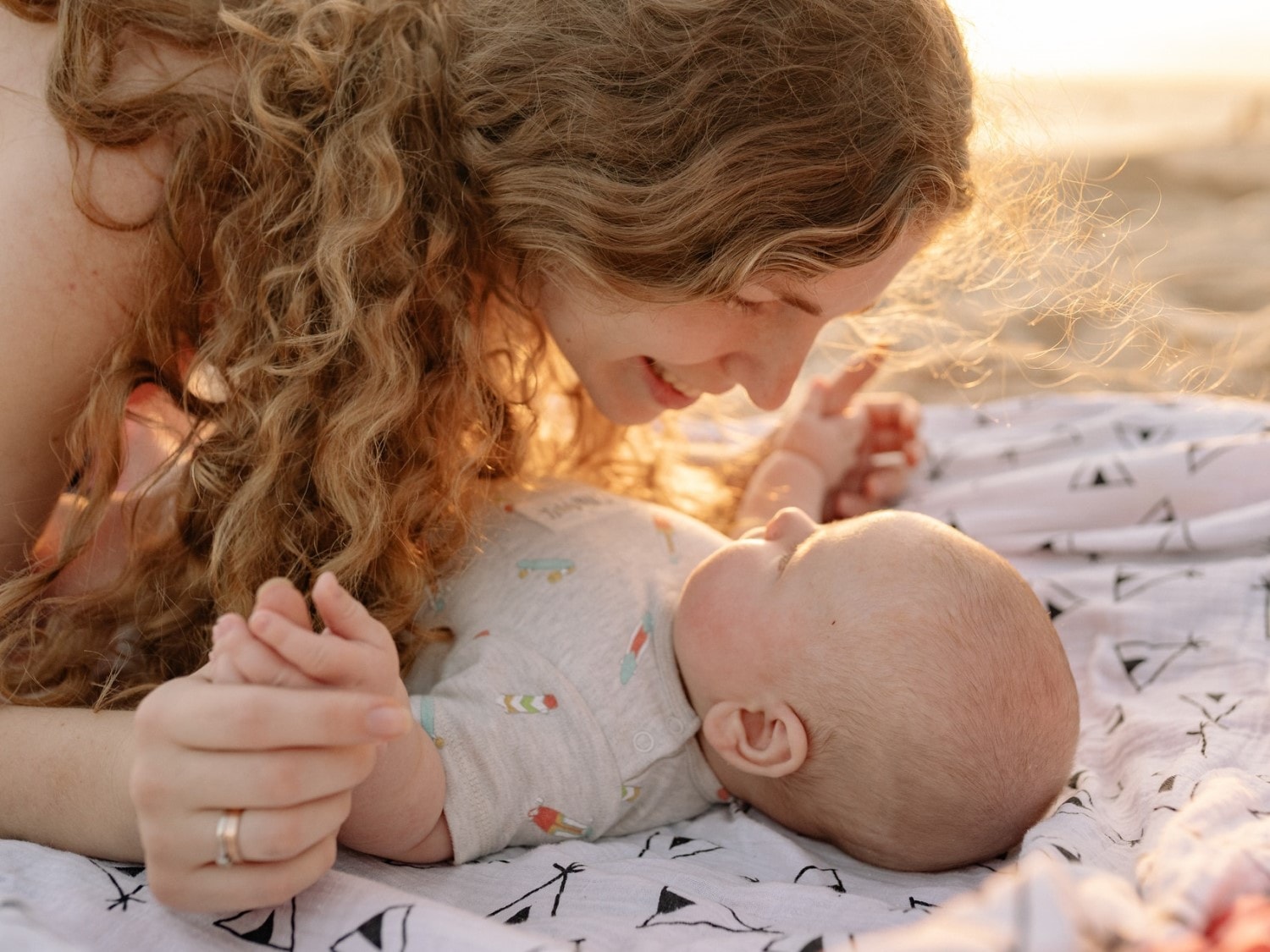 Eine Mutter mit lockigem Haar liegt auf einer Decke und hält die Hand ihres Babys, während sie es liebevoll ansieht.