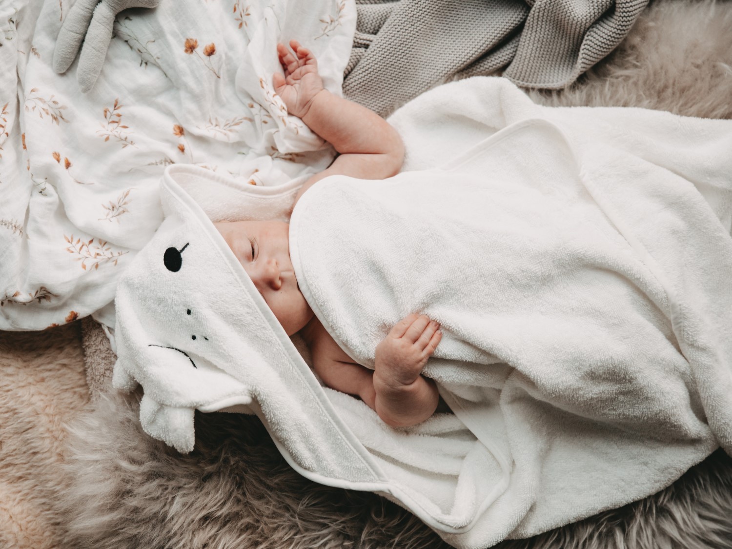 Ein schlafendes Baby, eingewickelt in ein weißes Handtuch mit Bärenohren, liegt auf einer weichen Decke mit Blumenmuster.
