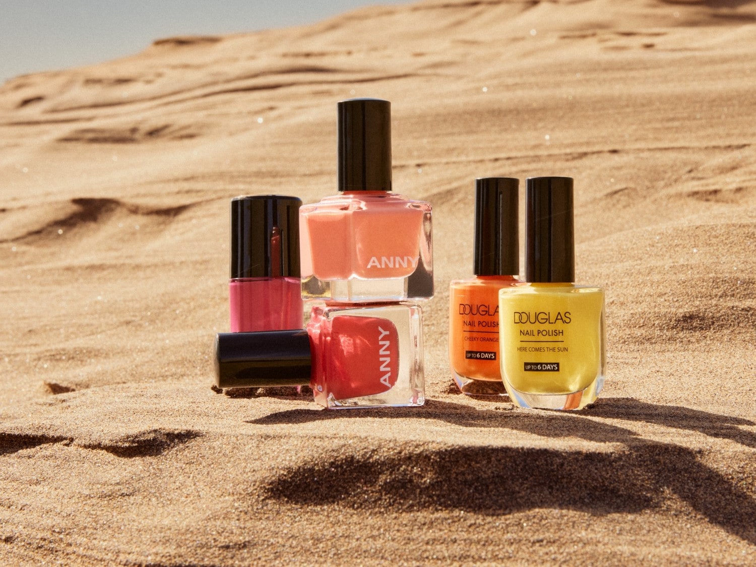 Verschillende kleuren nagellak van ANNY en Douglas, waaronder roze, oranje en geel, staan op een zandige ondergrond.