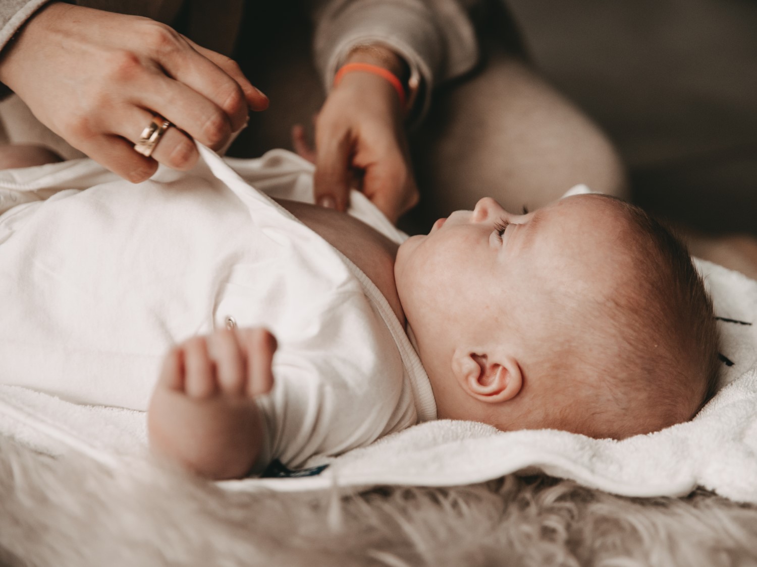 Ein Baby liegt auf einem weißen Handtuch, während eine Hand mit einem goldenen Ring ihm ein weißes Hemd anzieht.