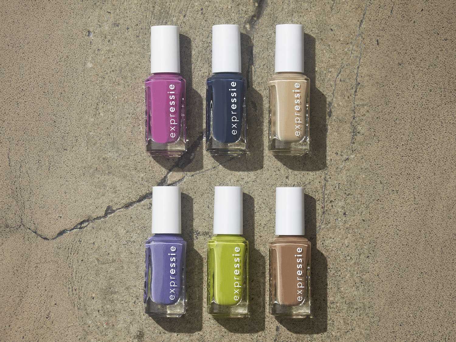 Seis frascos de esmalte de uñas Essie Expressie en tonos rosa, azul marino, beige, lila, verde lima y marrón claro, con tapas blancas, sobre una superficie de hormigón.