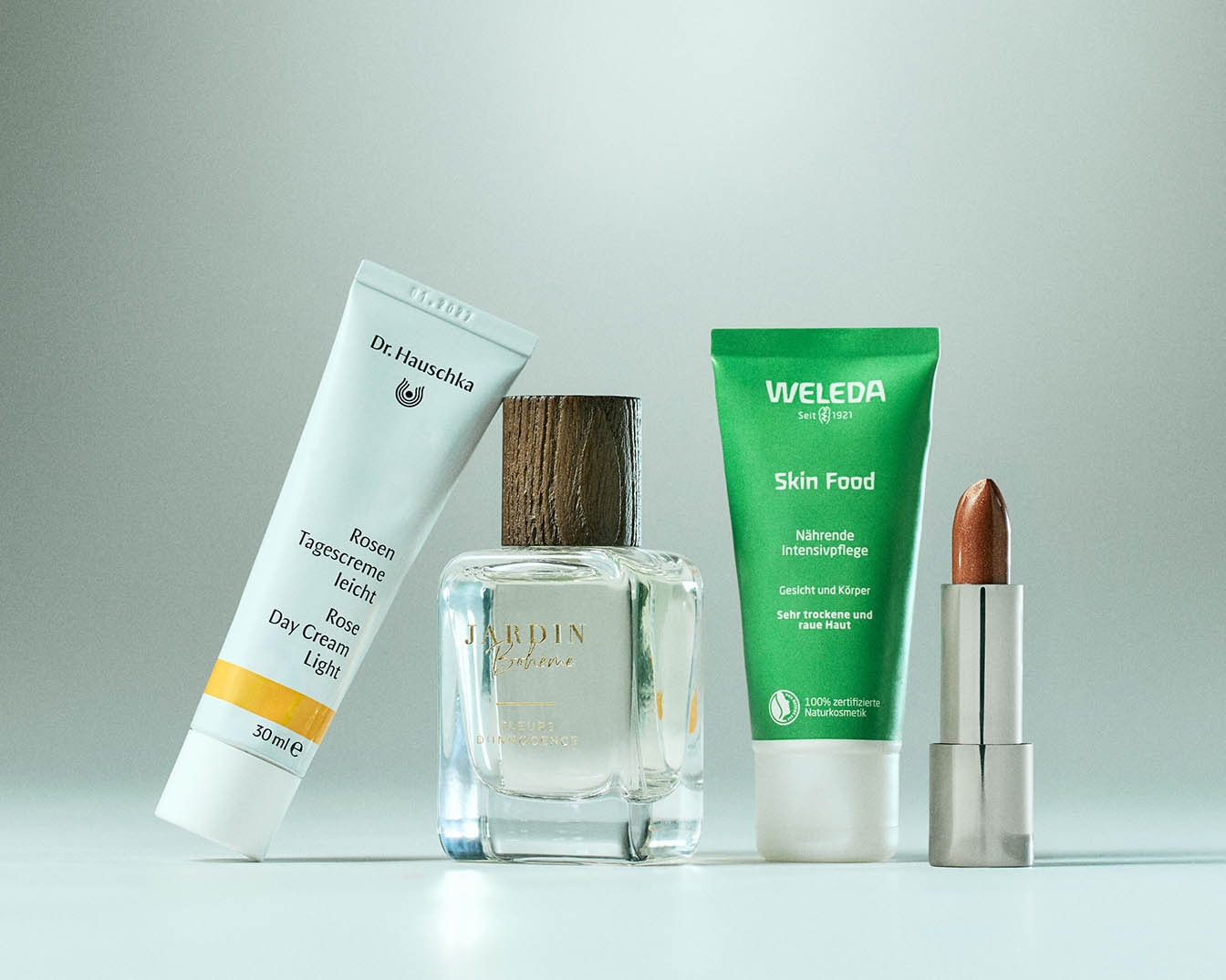 Na obrázku je pleťová kosmetika: Bílý krém Dr. Hauschka Rosen Tagescreme leicht, parfém Jardin Bohème, zelený krém Weleda Skin Food a bronzová rtěnka.