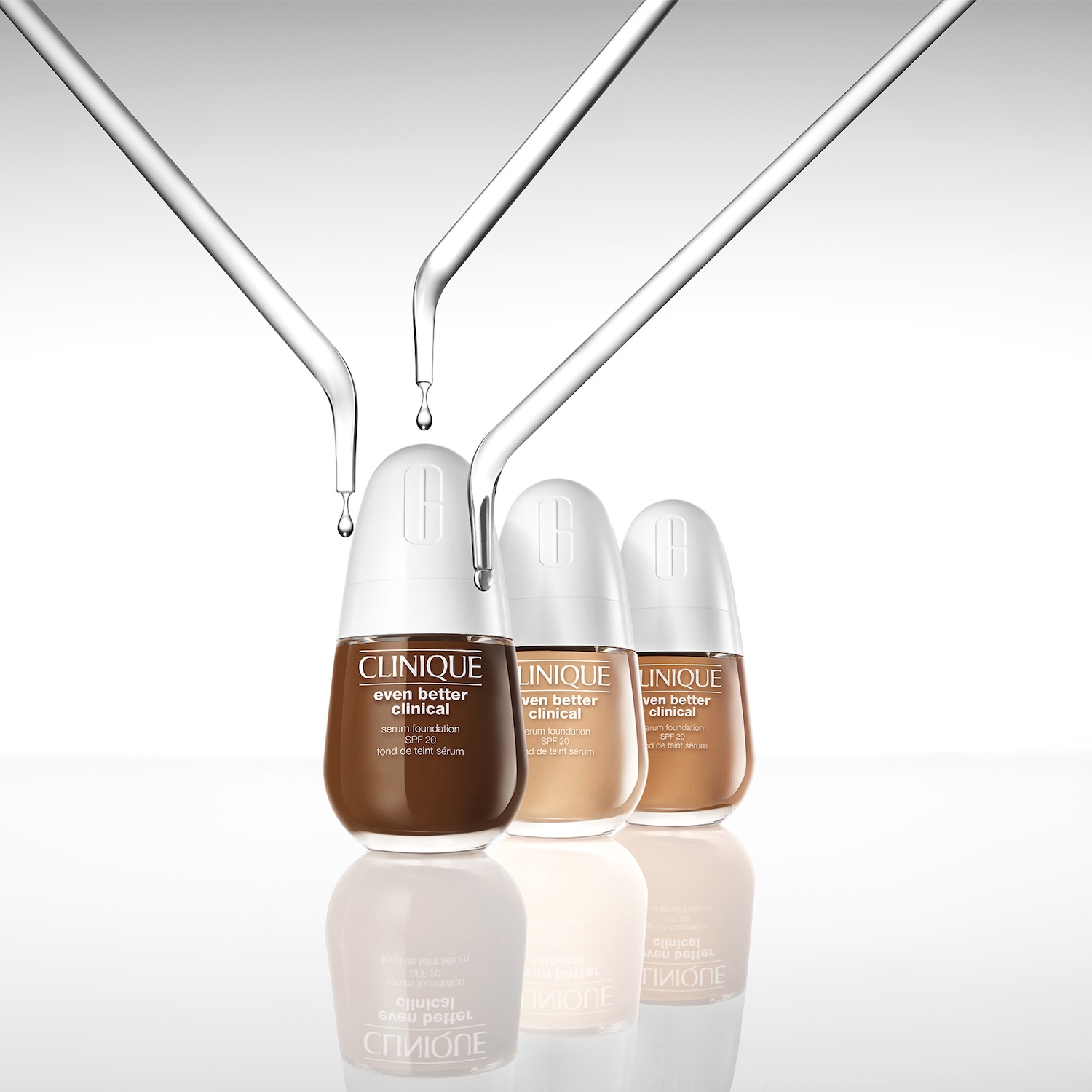 Drei Flaschen Clinique Even Better Clinical Serum Foundation SPF 20 in verschiedenen Farbtönen stehen auf einer reflektierenden Oberfläche, w ährend Serumtropfen von oben fallen.