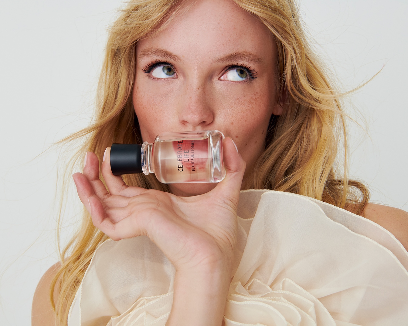 Blonďatá žena s pihami drží flakón parfému Celebrate Life od Beautiful Stories, s černým uzávěrem a průhlednou tekutinou.
