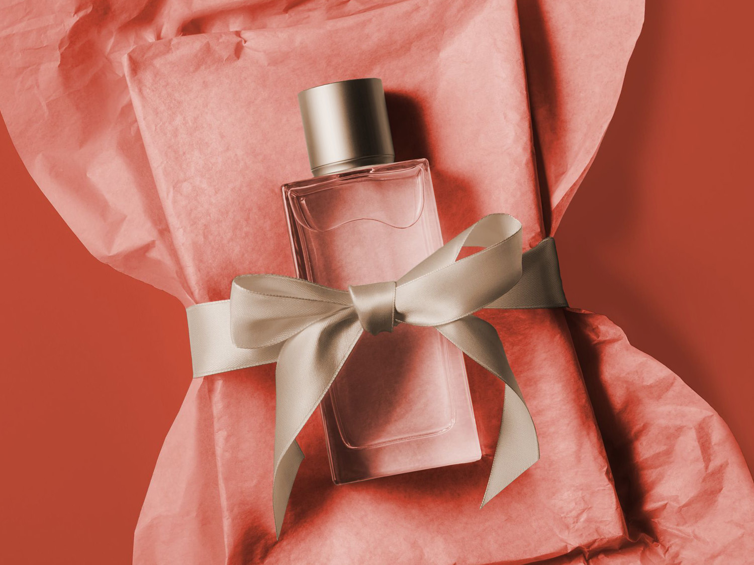 Un flacon de parfum rectangulaire avec un bouchon argenté est posé sur du papier de soie froissé de couleur corail, noué avec un ruban de satin beige, créant un cadeau élégant.