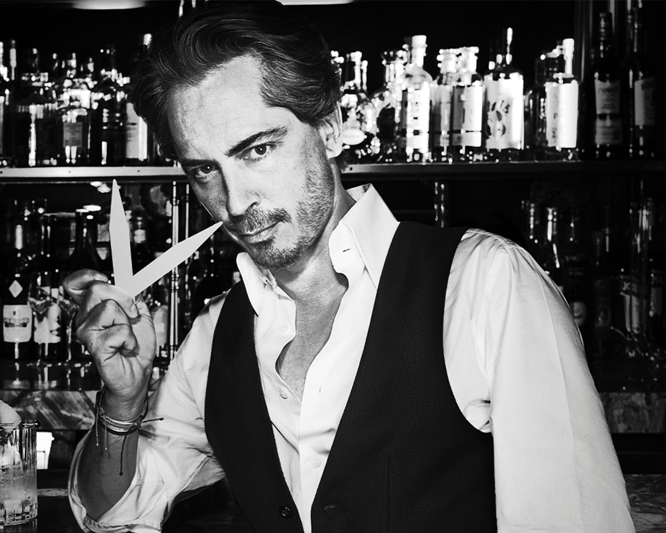 Čiernobiela fotografia muža v košeli a veste, ktorý drží papierový prúžok pod nosom, s barom v pozadí.