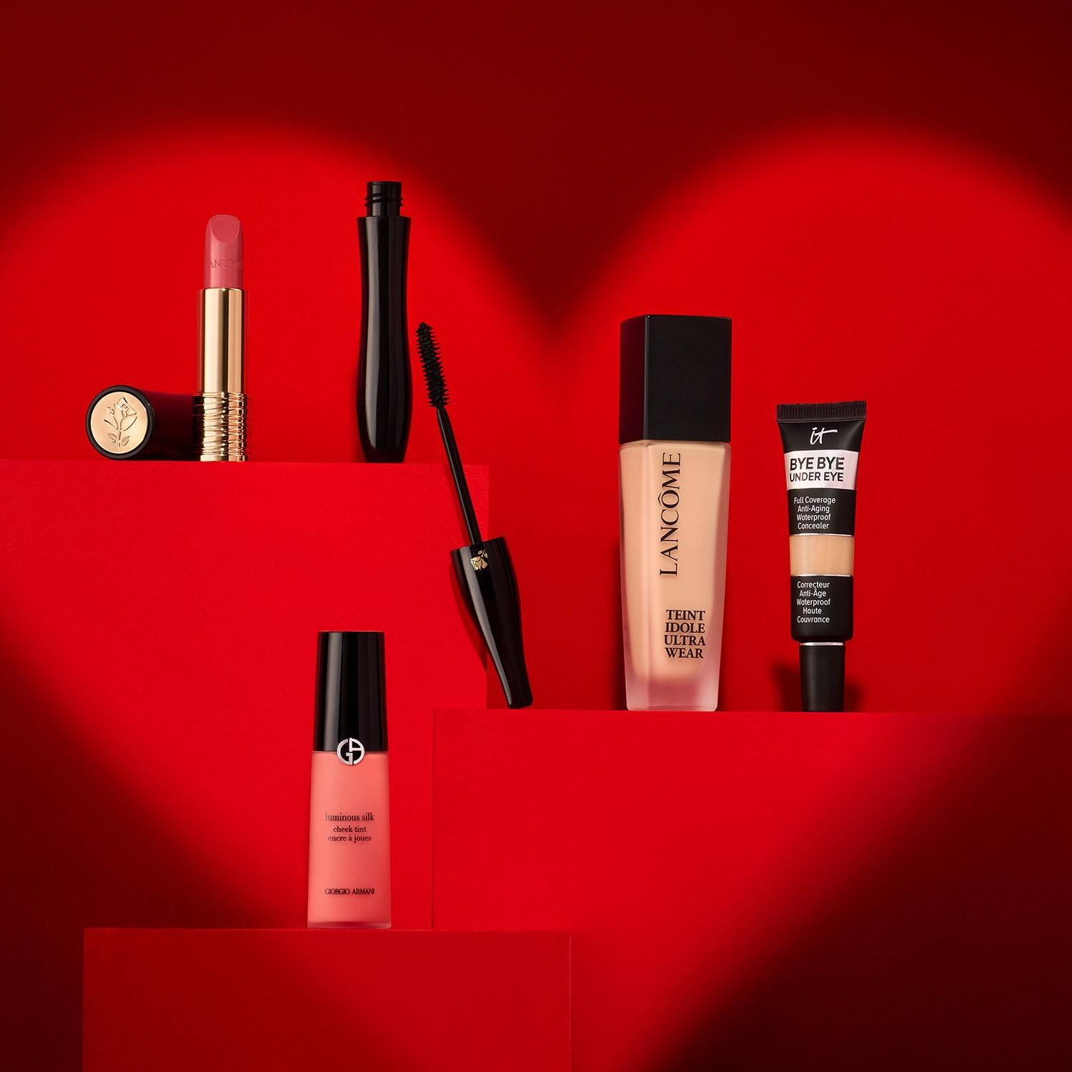 Découvrez un assortiment de produits de beauté, comprenant un rouge à lèvres doré, un mascara noir, un fond de teint Lancôme, un correcteur anti-âge et un blush Giorgio Armani, le tout sur un fond rouge en forme de cœur.