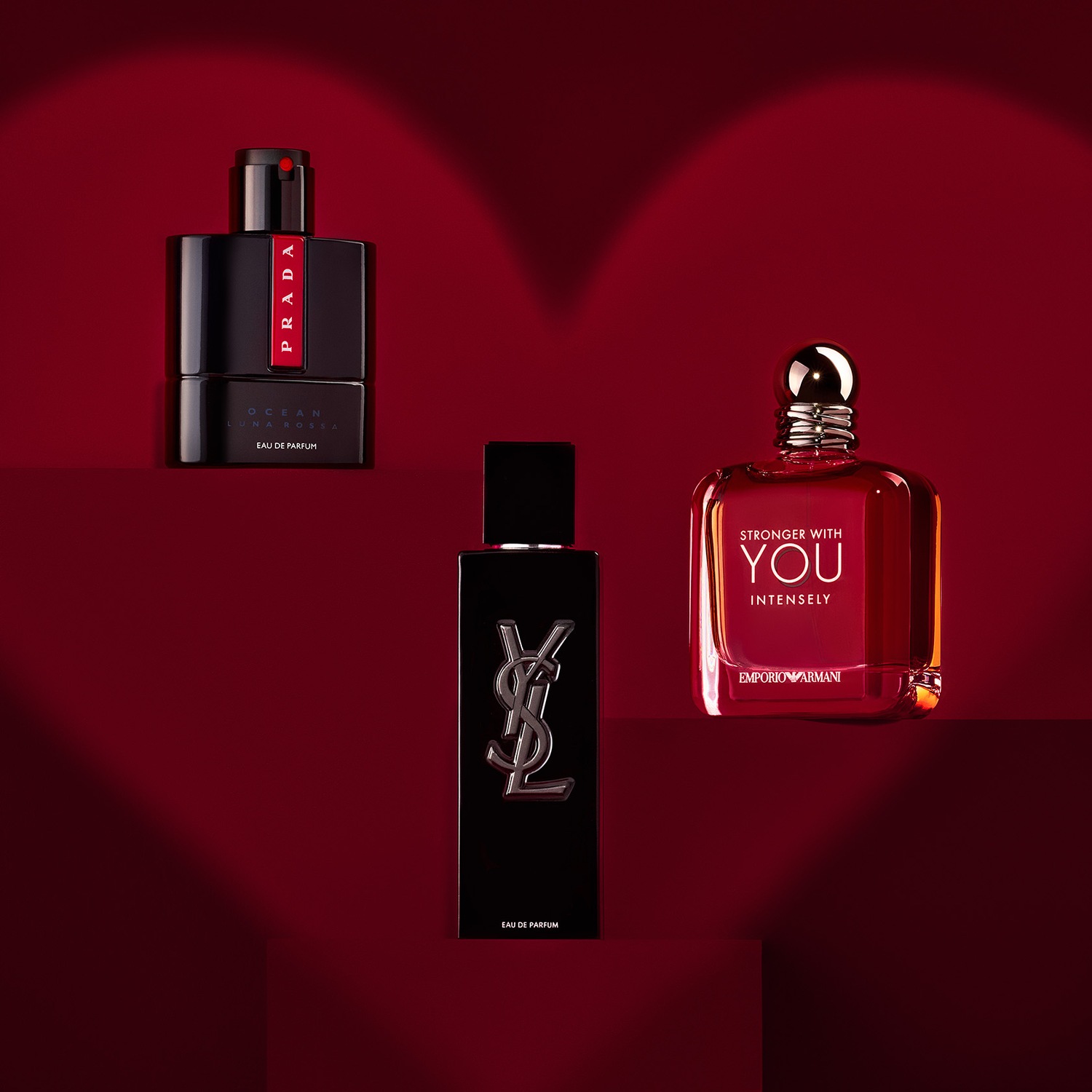 Découvrez un trio de parfums masculins présentés sur un fond rouge foncé, comprenant Prada Ocean Luna Rossa, YSL et Emporio Armani Stronger With You Intensely.