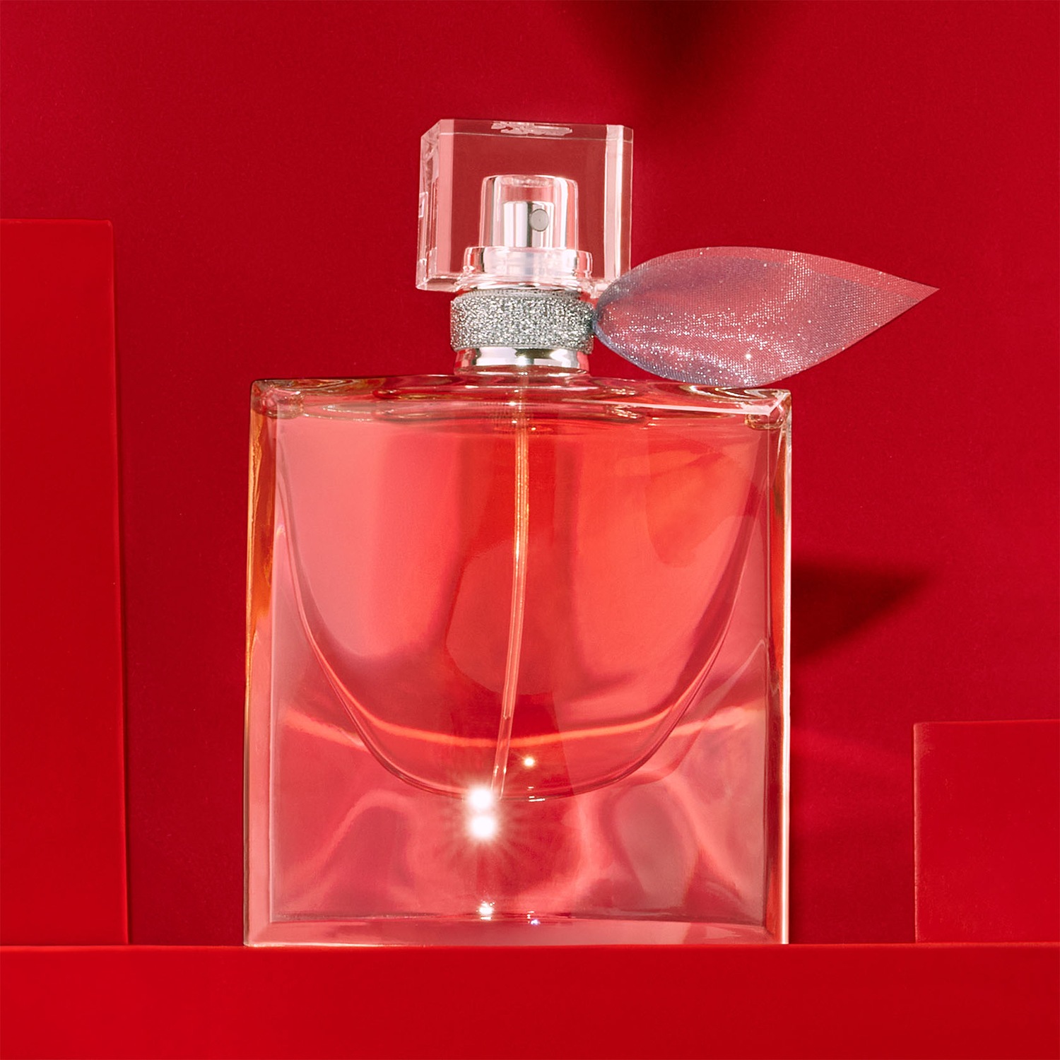Un flacon de parfum en verre transparent rempli d'un liquide rose, orné d'un ruban argenté scintillant, se dresse sur un fond rouge assorti, évoquant l'élégance et le luxe.