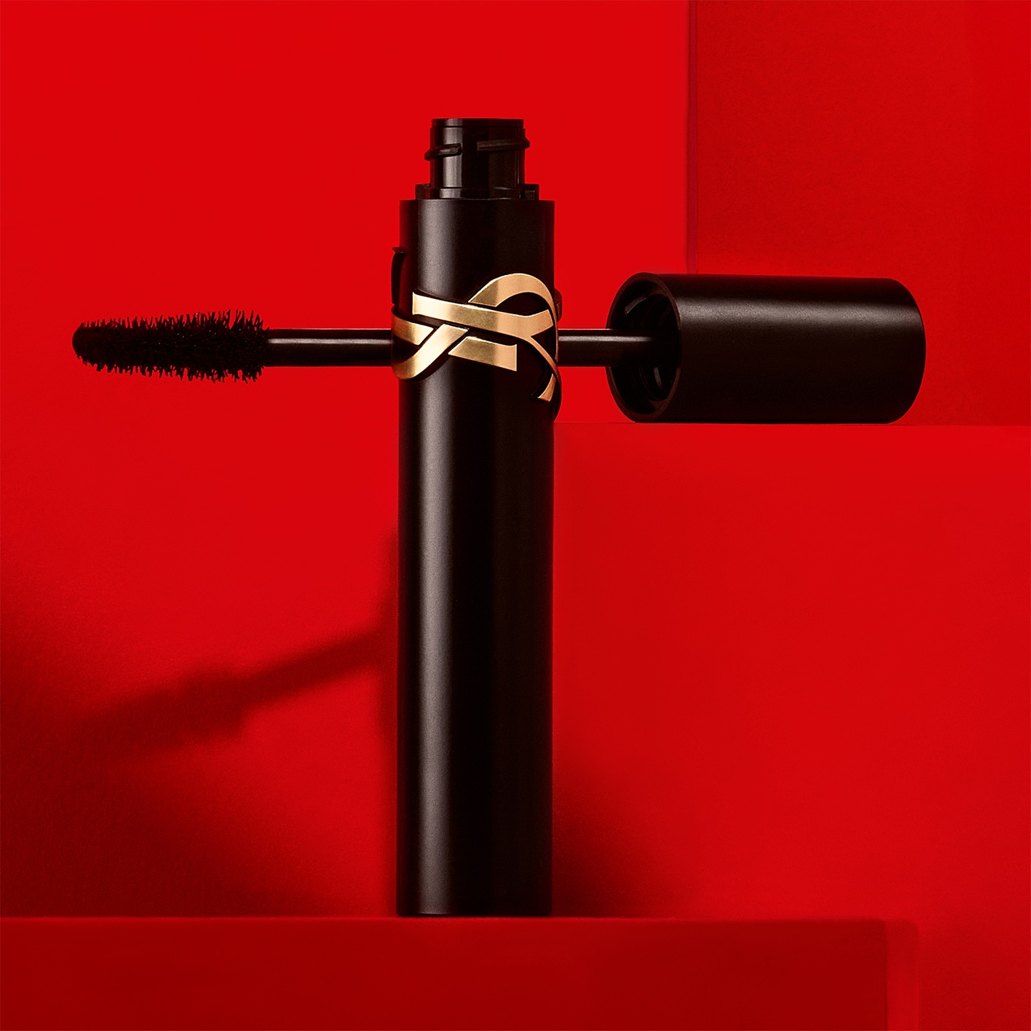 Voici un mascara noir élégant de Yves Saint Laurent, orné du logo doré emblématique, avec une brosse visible, le tout sur un fond rouge vibrant.