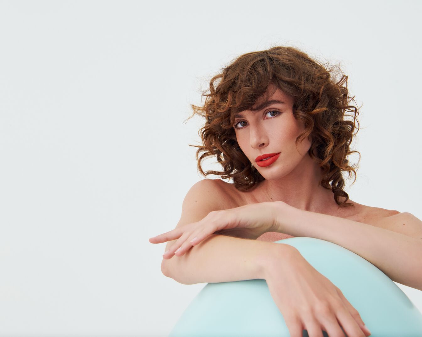 Porträt einer Frau mit lockigem, braunem Haar und roten Lippen, die auf einem hellblauen Stuhl sitzt und in die Kamera blickt.