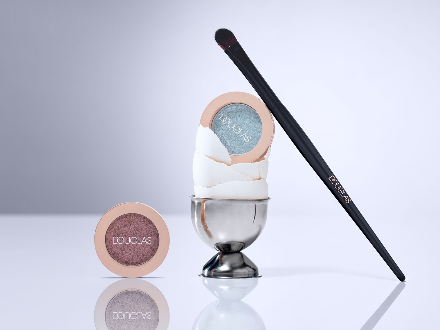 Nahaufnahme von Douglas Lidschatten in einem silbernen Eierbecher mit einem schwarzen Pinsel, der sich auf einem glänzenden, reflektierenden Tisch befindet.