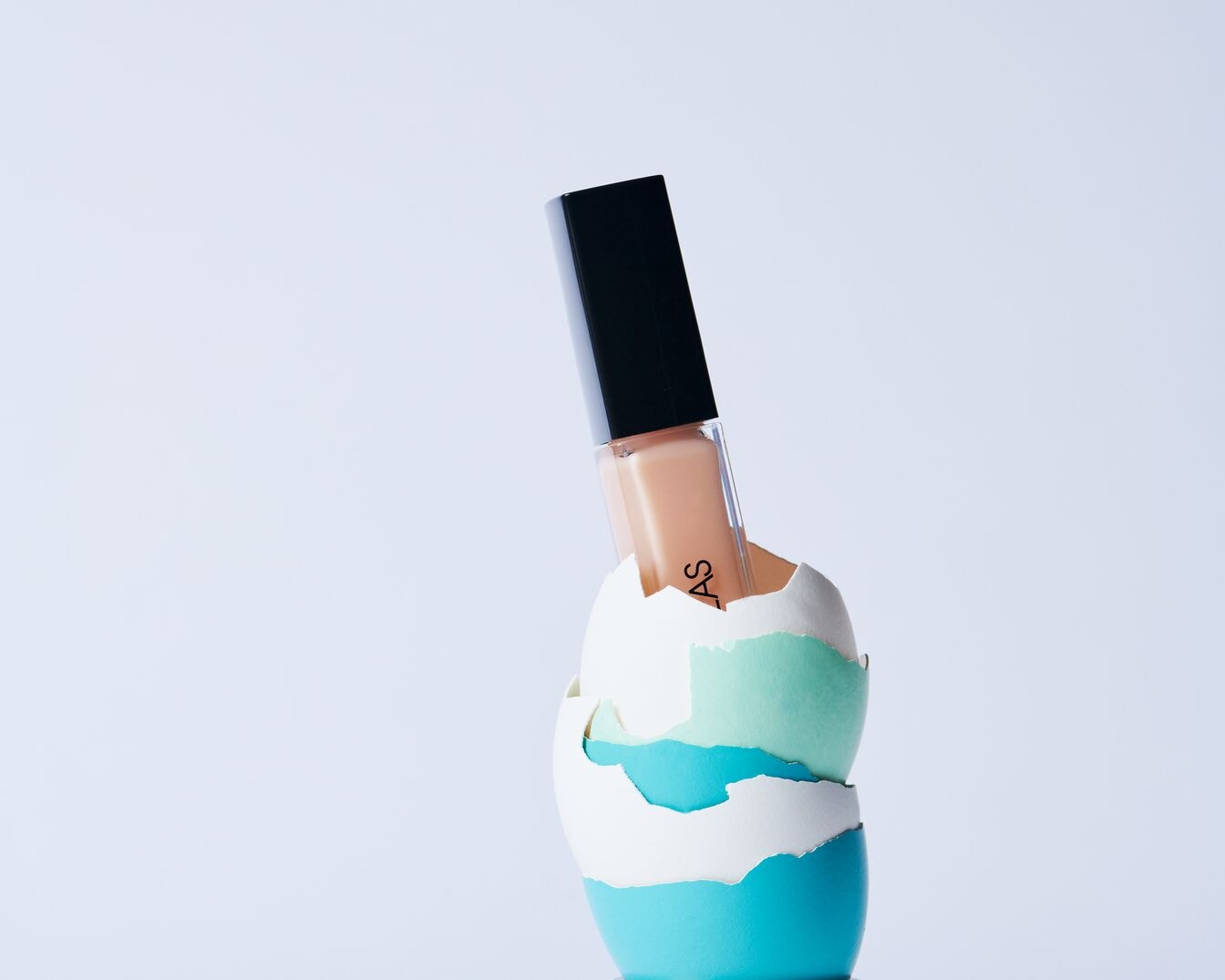 Fondotinta color pesca Douglas con tappo nero, esposto in un guscio d'uovo decorativo a strati bianchi, verdi e blu.