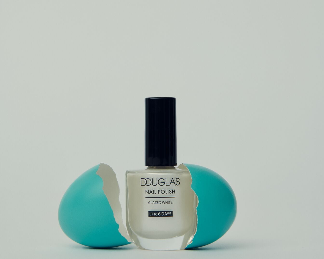 Smalto per unghie Douglas "Bianco Glacé" in flacone trasparente con tappo nero, presentato all'interno di un guscio d'uovo turchese rotto, per unghie perfette fino a 6 giorni.