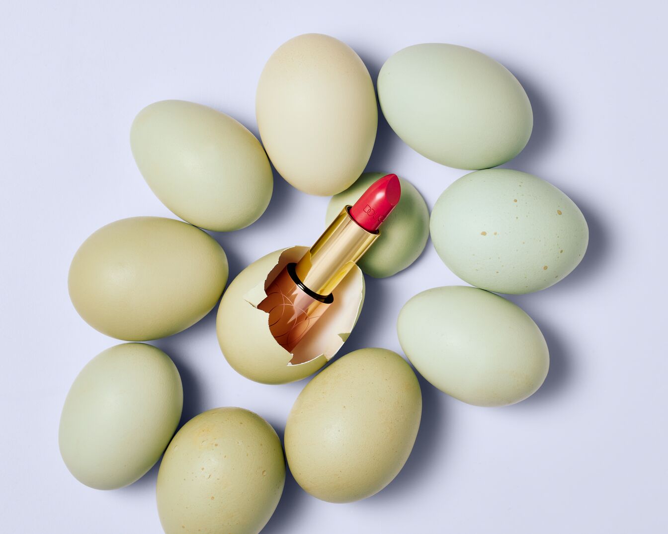 Rossetto rosso vibrante emerge da un guscio d'uovo rotto, circondato da uova color pastello, simbolo di bellezza che sboccia in primavera.