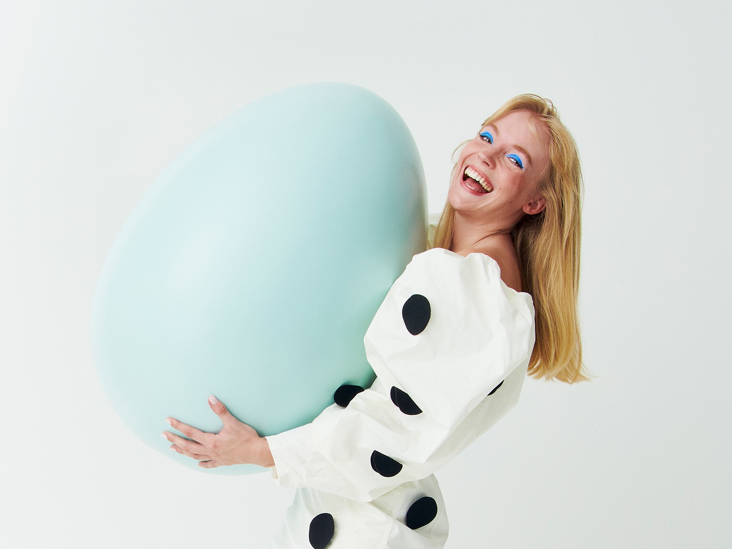 Una mujer rubia sonríe alegremente mientras sostiene un huevo azul gigante, con maquillaje azul brillante en los ojos y vestida con una blusa blanca con lunares negros.