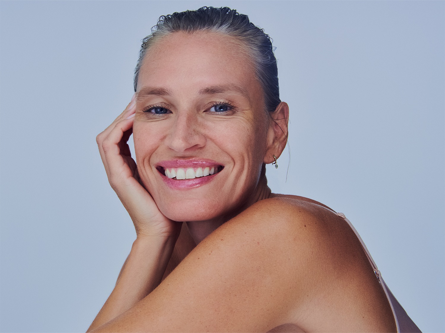 Portrait d'une femme souriante aux cheveux gris tirés en arrière, la main sur le visage, avec un fond bleu clair.