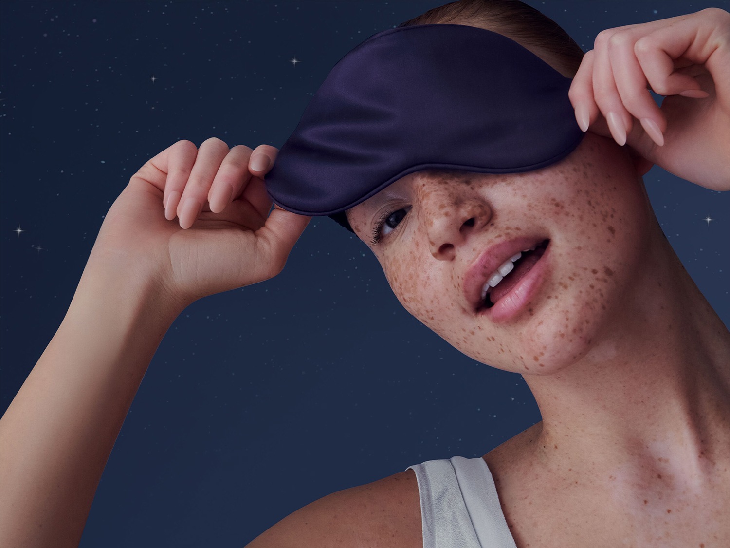 Une femme aux cheveux tirés en arrière, portant un masque de sommeil bleu foncé sur les yeux, se prépare pour une nuit de sommeil paisible.