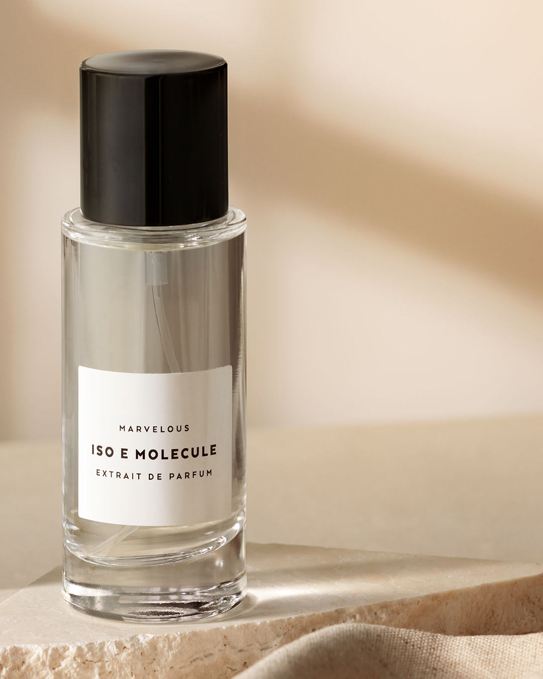 Przezroczysta butelka perfum Marvellous Iso E Molecule Extrait de Parfum z czarną zakrętką na teksturowanym beżowym tle.