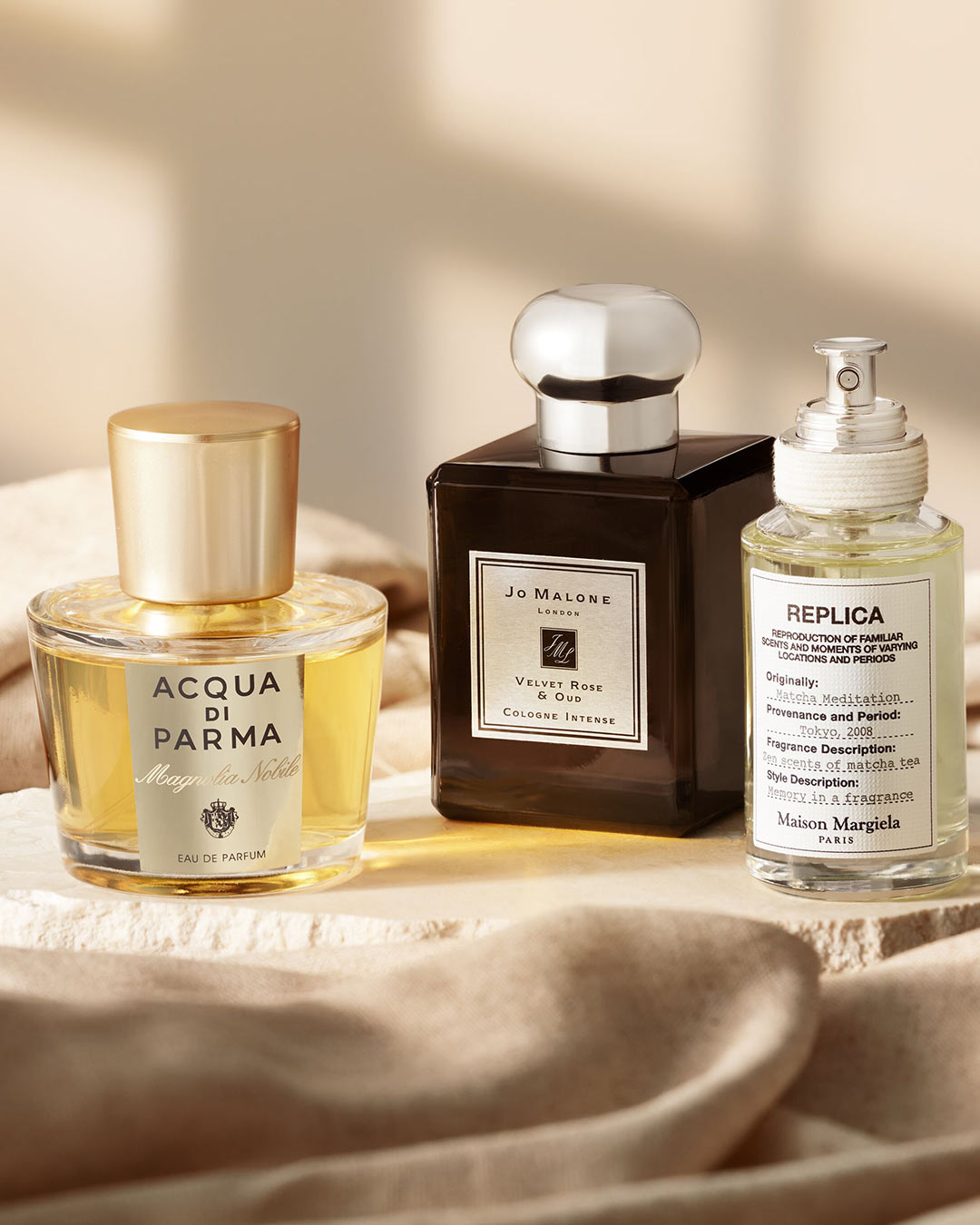 Elegancka kompozycja trzech flakonów perfum: Acqua di Parma Magnolia Nobile o złotym odcieniu, Jo Malone Velvet Rose & Oud w ciemnobrązowym flakonie oraz Replica Matcha Meditation od Maison Margiela.