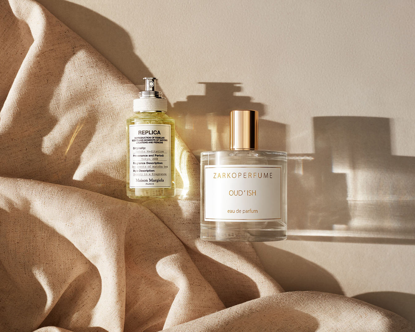Dwa flakony perfum: "REPLICA Matcha Meditation" o jasnożółtym zabarwieniu i "ZARKOPERFUME OUD'ISH" z przezroczystego szkła ze złotą nakrętką, oba na tle beżowej tkaniny.