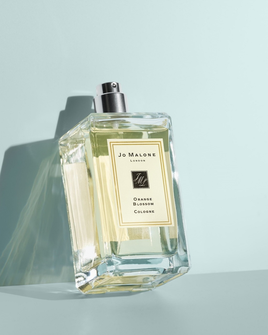 Ein rechteckiger, transparenter Glasflakon von Jo Malone London Orange Blossom Cologne mit silbernem Sprühkopf, gefüllt mit einer hellgelben Flüssigkeit, steht auf einer hellblauen Oberfläche.