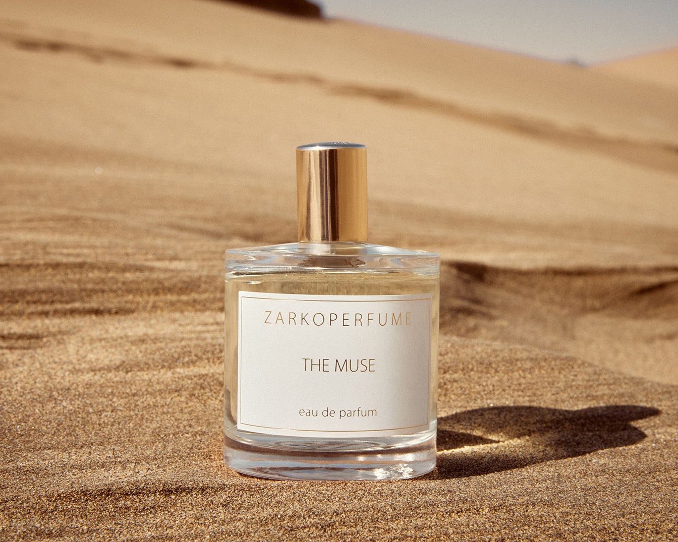 El frasco de perfume transparente "The Muse" de Zarkoperfume, con tapón dorado, destaca sobre la arena dorada, evocando una sensación de lujo y calidez.