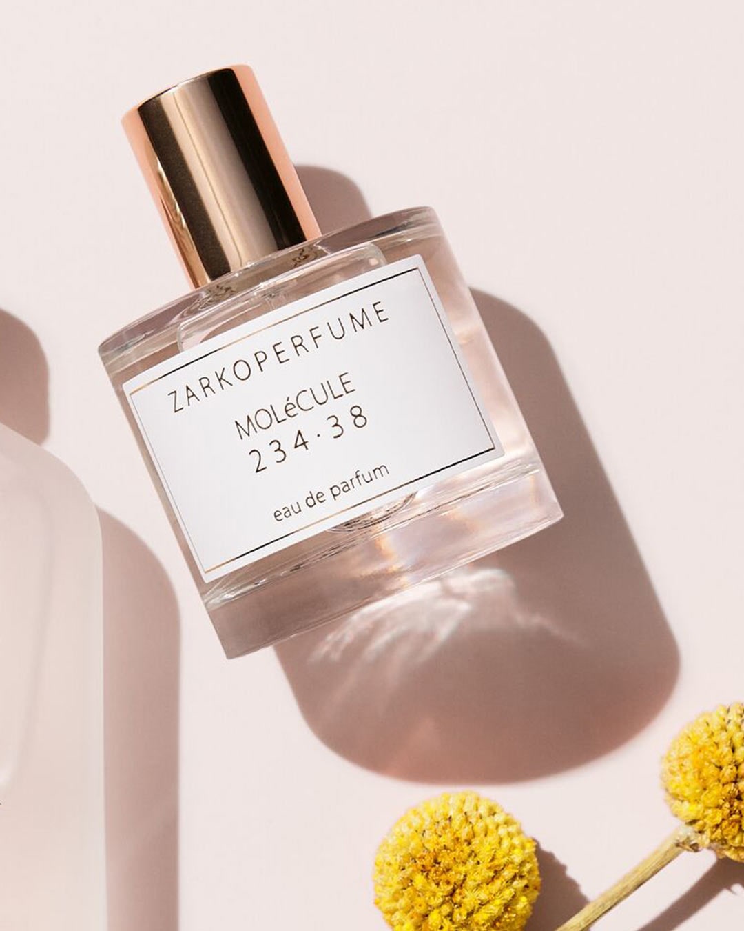 El frasco de perfume ZARKOPERFUME Molécule 234.38 Eau de Parfum tiene una tapa dorada y una etiqueta blanca con letras negras, sobre un fondo rosa pálido con flores amarillas.