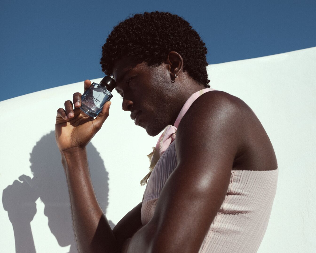 Un homme afro-américain tient un flacon de parfum bleu clair "JOOP!" devant un mur blanc sous un ciel bleu, évoquant une sensation de fraîcheur et de sophistication.