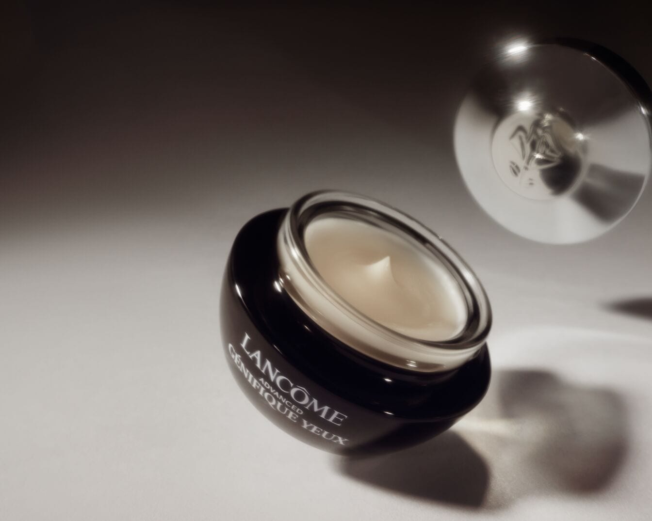 Nahaufnahme einer geöffneten schwarzen Dose Lancôme Génifique Yeux Augencreme, die eine cremefarbene Creme zeigt, mit einem silbernen Deckel im Hintergrund.