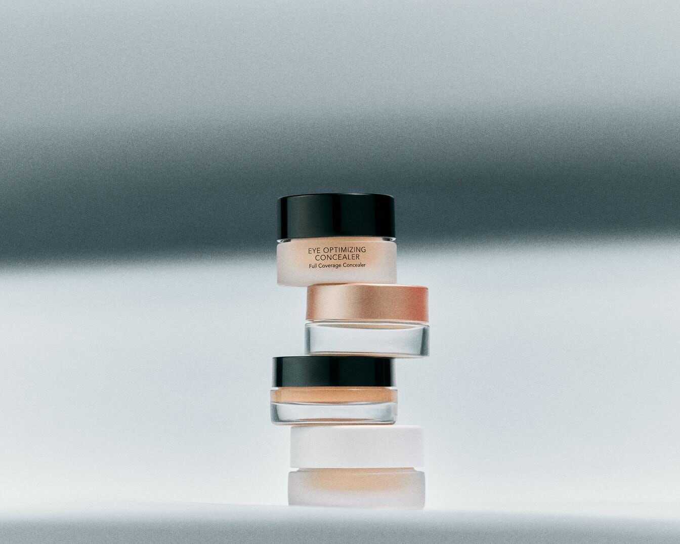 Štai bokštas iš „Eye Optimizing Concealer“ indelių su juodais dangteliais, skirtų pilnam maskavimui, sukrautas ant pilko paviršiaus.