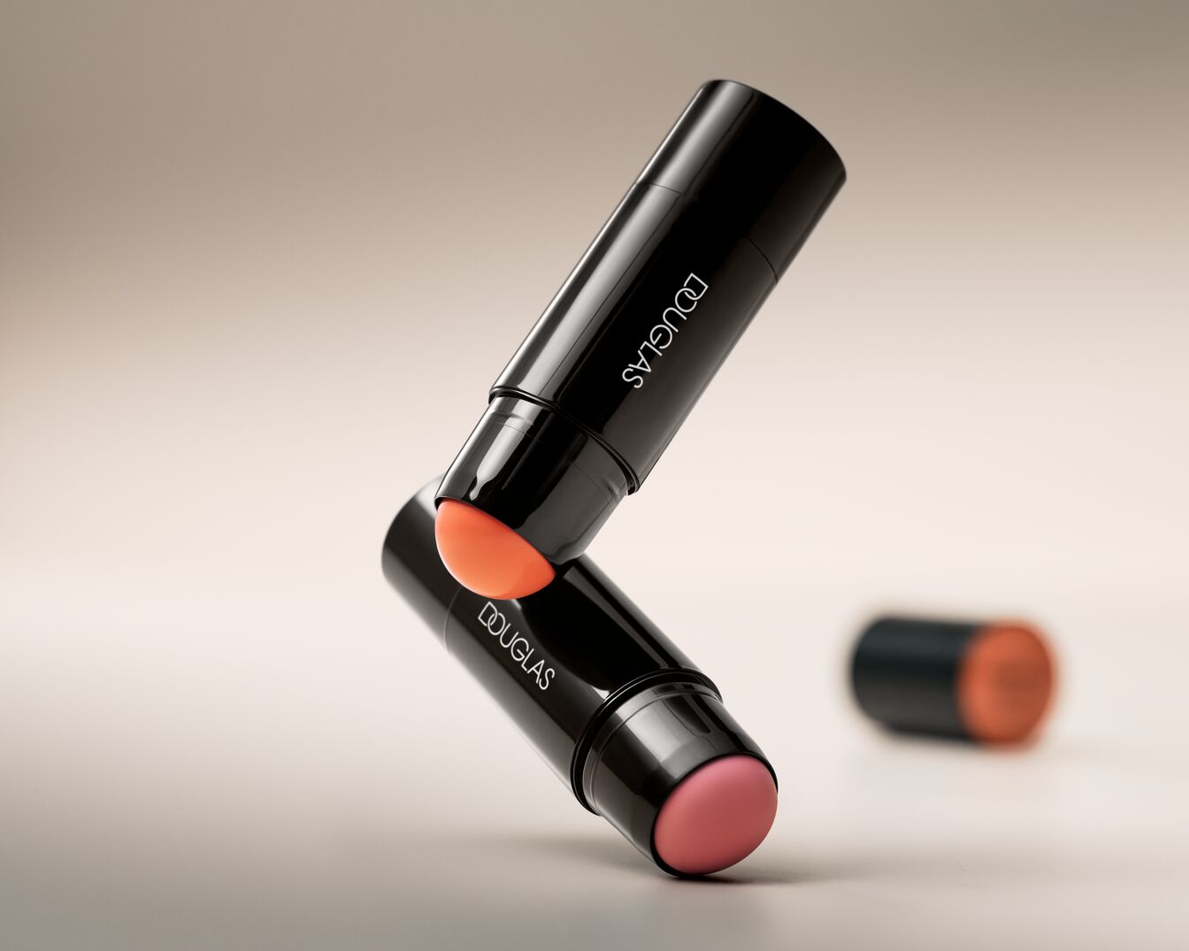 Dwa błyszczące sztyfty do makijażu Douglas w czarnych opakowaniach, jeden z pomarańczowym, a drugi z różowym odcieniem, ułożone jeden na drugim.
