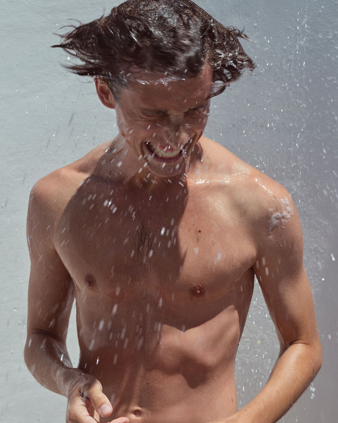 Jaunas vyras be marškinių, besimaudantis po dušu, su šlapiais plaukais ir putomis ant kūno, besišypsantis užmerktomis akimis.