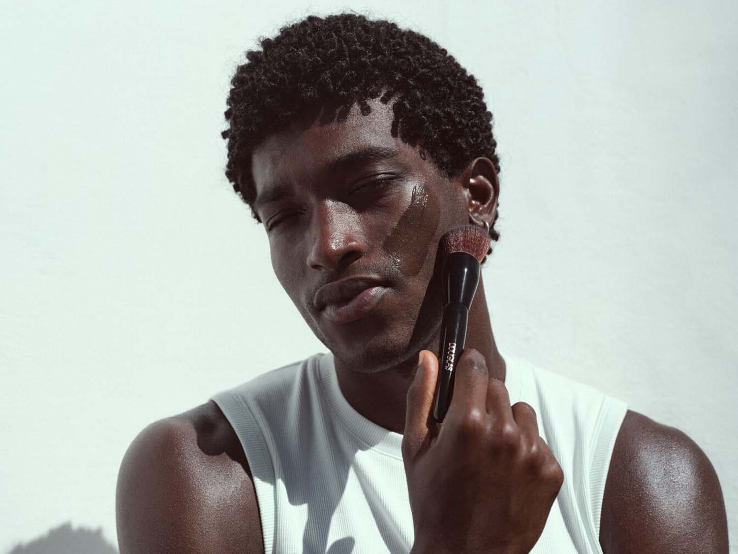 Tamsiaodis vyras tepa veidą rudu skysčiu su juodu makiažo šepetėliu, vilkėdamas baltą berankovę palaidinę.