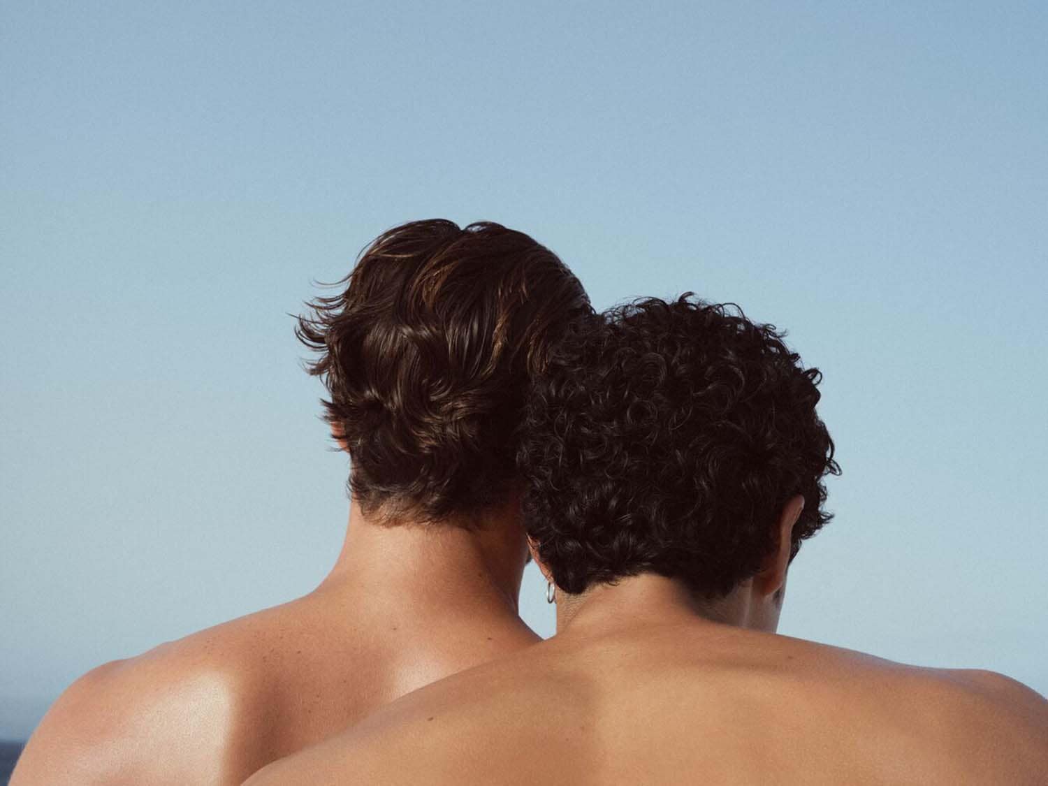 Du vyrai, stovintys nugara į kamerą, vienas su tiesiais, o kitas su garbanotais plaukais, priešais mėlyną dangų.