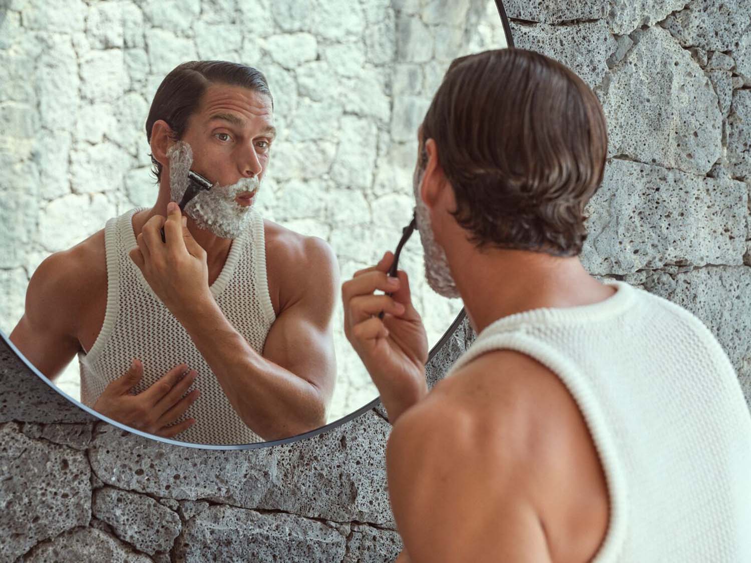 Vyras su baltu trikotažiniu viršumi skuta barzdą skustuvu priešais veidrodį, demonstruodamas asmens priežiūros rutiną.