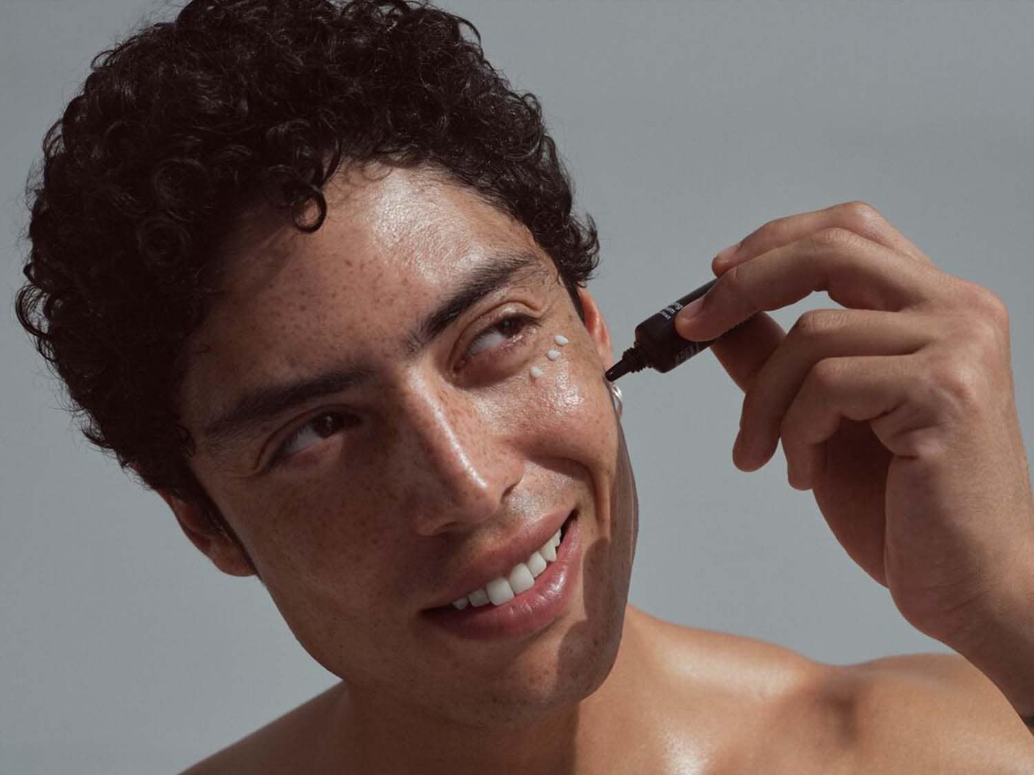 Jaunuolis tepa paakių kremą iš juodo tūbelės, šypsodamasis ir žiūrėdamas į viršų, kad oda būtų sveika.