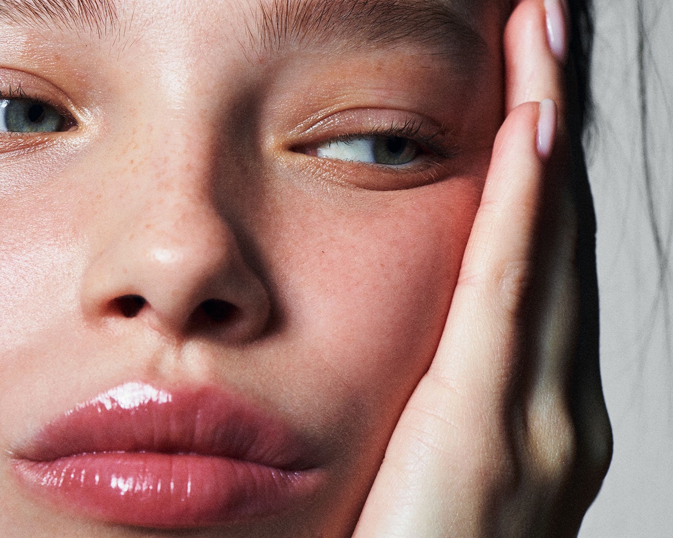 Nahaufnahme eines jungen Gesichts mit Sommersprossen, glänzenden Lippen und einer Hand, die die Wange berührt.