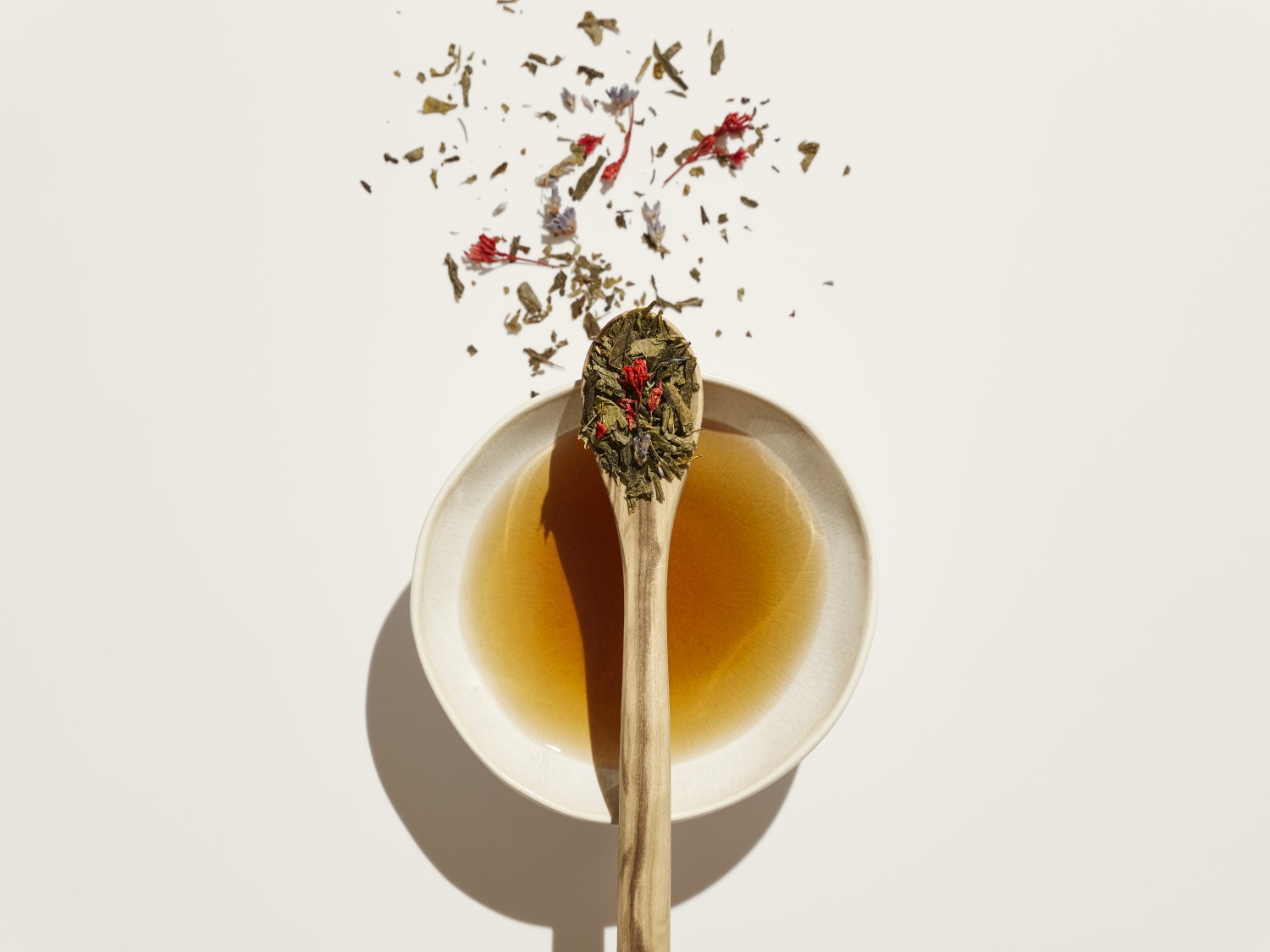 Draufsicht auf eine Schale mit Tee, einem Holzlöffel voller grüner Teeblätter und roten Blüten auf einem weißen Hintergrund.