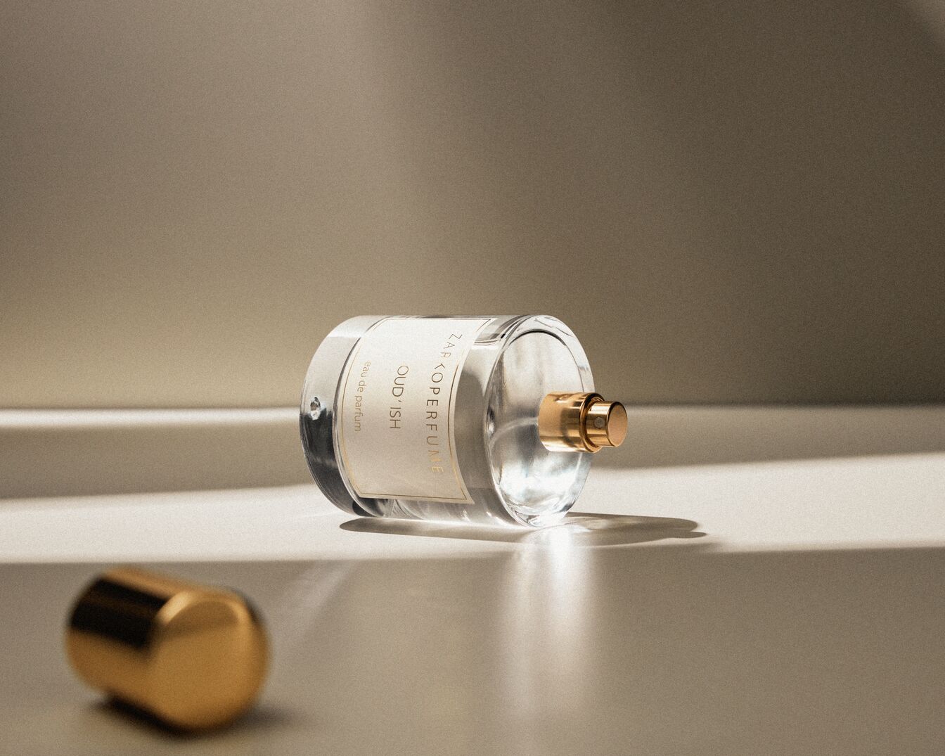 Eine durchsichtige Glasflasche Zarkoperfume Oud'ish Eau de Parfum liegt auf der Seite, die goldene Kappe daneben.