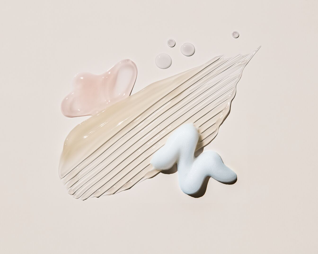 Švelniai rožinio, kreminio ir mėlyno atspalvių kosmetikos produktų tekstūros ant šviesaus fono.