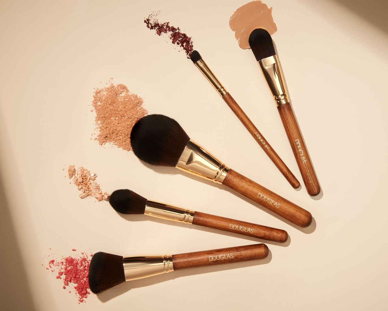 Verschiedene Make-up-Pinsel mit Holzgriffen und goldenen Akzenten sind auf einem hellen Hintergrund angeordnet, umgeben von Farbpigmenten in Rosa-, Braun- und Beigetönen.