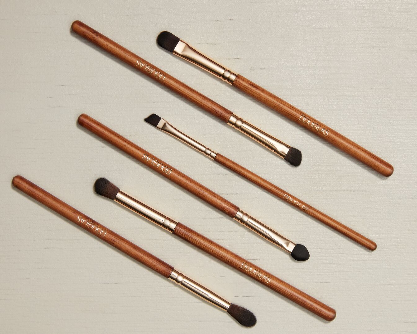 Eine Nahaufnahme von Douglas Make-up-Pinsel-Set mit Holzgriffen und goldenen Akzenten, die auf einem hellen Hintergrund angeordnet sind.