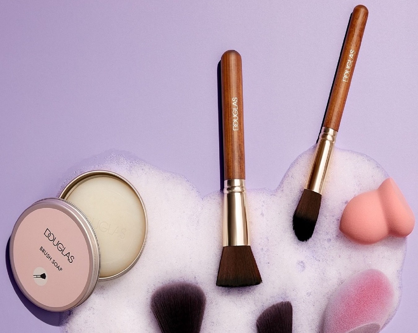 Auf einem lila Untergrund liegen Make-up-Pinsel mit Holzgriffen, ein rosa Schwämmchen und eine Dose Douglas Brush Soap in einer weißen Schaumwolke.
