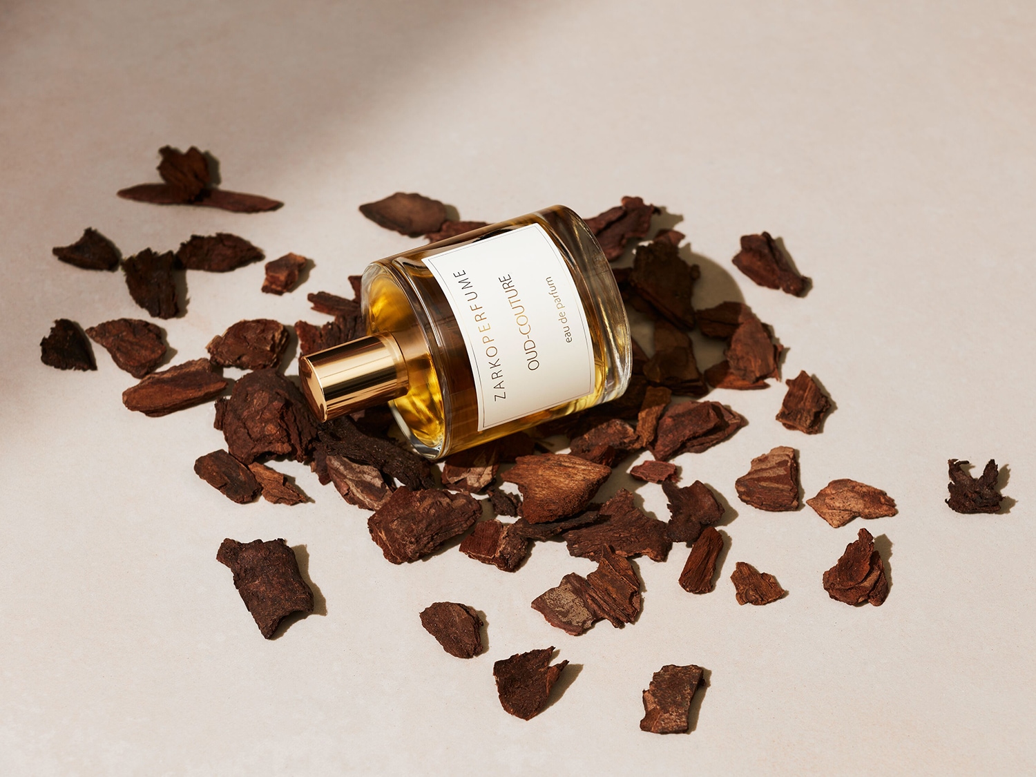 Ein klares Glasflakon Zarkoperfume Oud-Couture Eau de Parfum liegt auf einer hellen Oberfläche, umgeben von kleinen, dunklen Holzstücken.