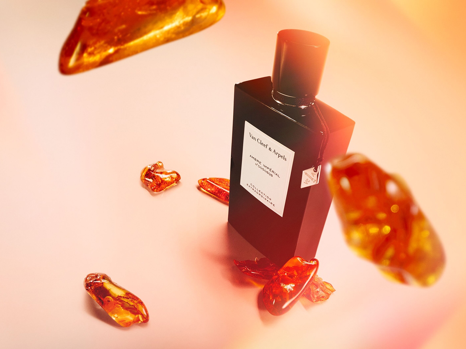 Frasco negro de perfume Van Cleef & Arpels Ambre Impérial rodeado de trozos de ámbar sobre un fondo rosa anaranjado.