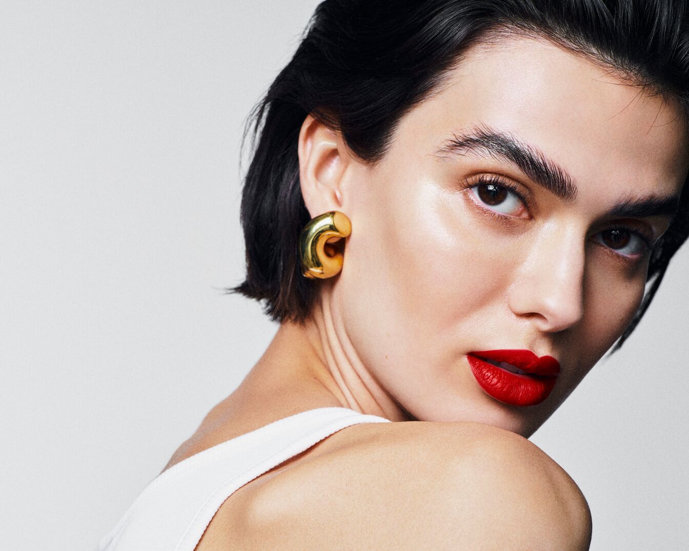 Nahaufnahme einer Frau mit roten Lippen, goldenen Ohrringen und zurückgekämmtem, dunklem Haar, die über die Schulter in die Kamera blickt.