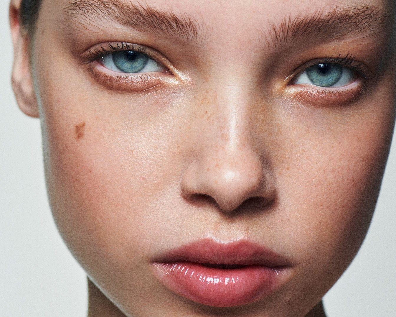 Nahaufnahme eines jungen Gesichts mit hellblauen Augen, Sommersprossen und einem Muttermal auf der Wange, die Lippen sind glänzend hervorgehoben.