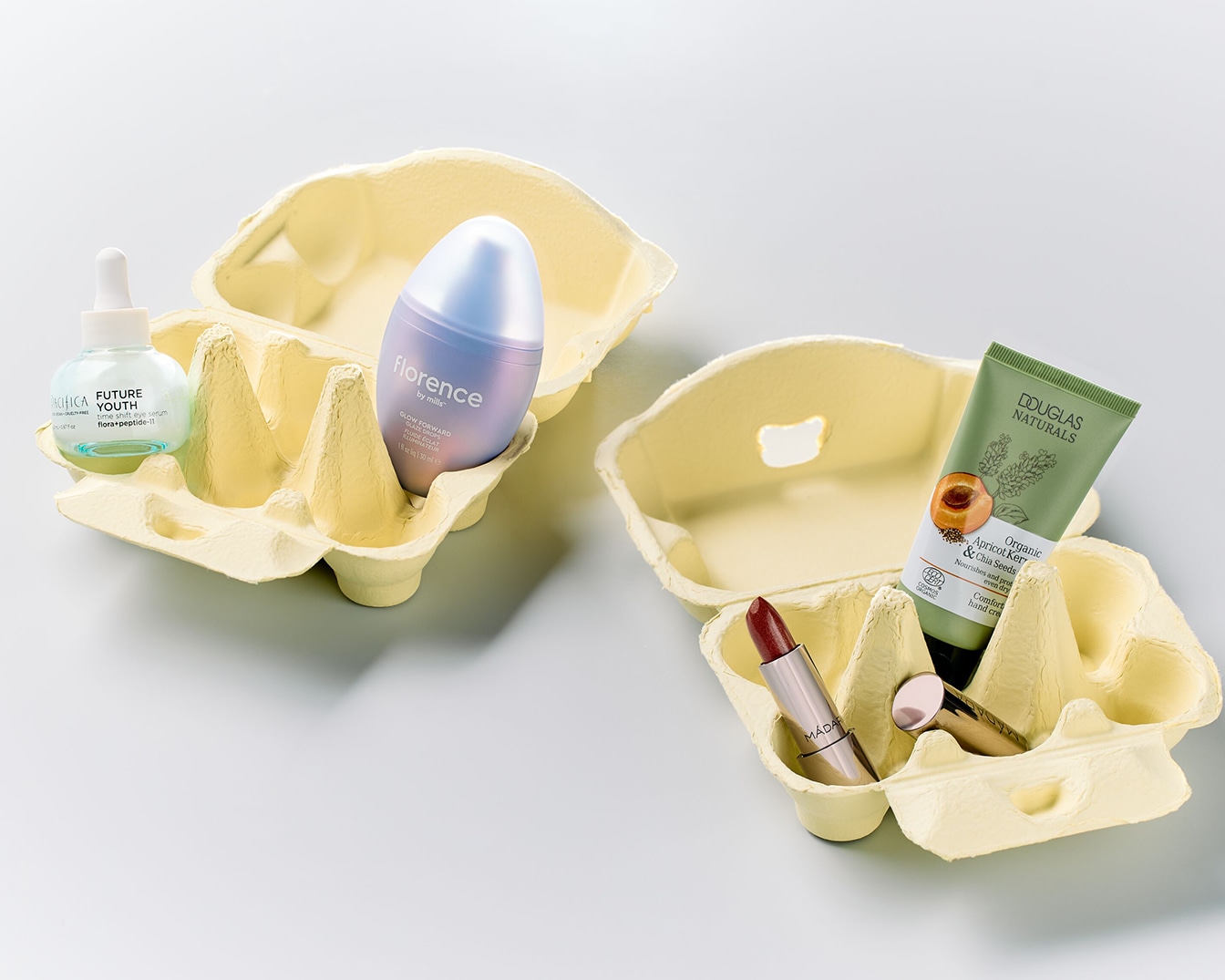 Dos hueveras de cartón con productos de belleza: un sérum en frasco de cristal con pipeta, una crema hidratante en envase lila, un labial burdeos, una crema de manos en tubo verde.