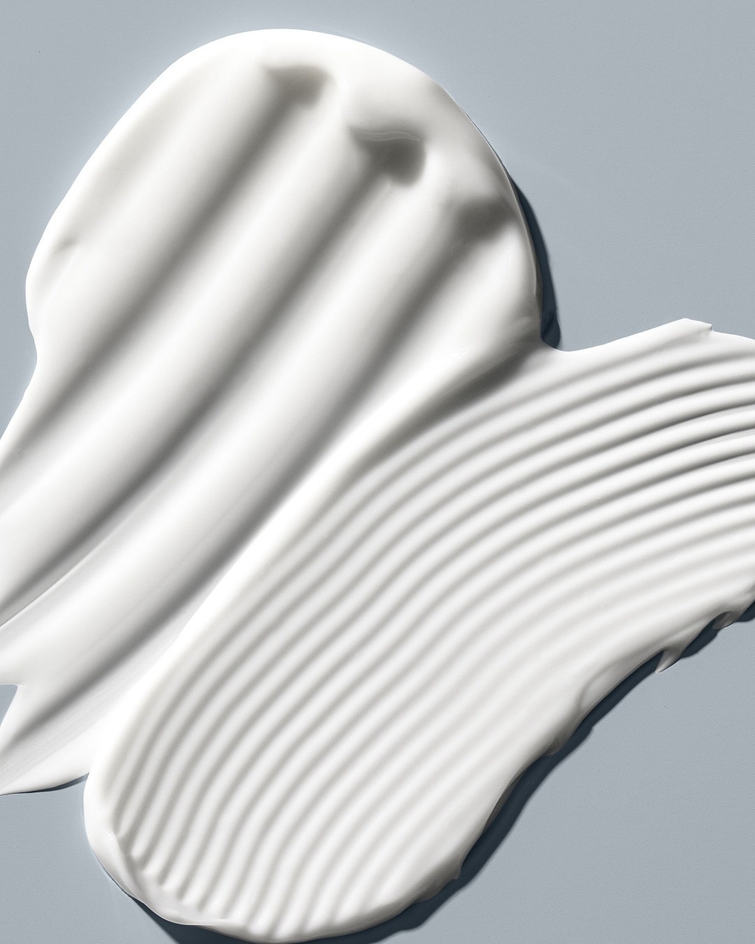 Primer plano de una muestra de crema blanca sobre un fondo azul claro, mostrando una textura rica y ondulada, ideal para hidratar y suavizar la piel.